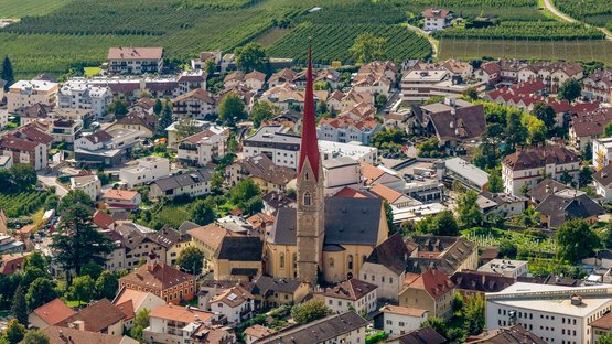 Hotels in Schlanders: erstklassiger Urlaub Luftaufnahme einer Stadt mit Kirche und roten Kirchturmspitze umgeben von Wohngebäuden und Feldern